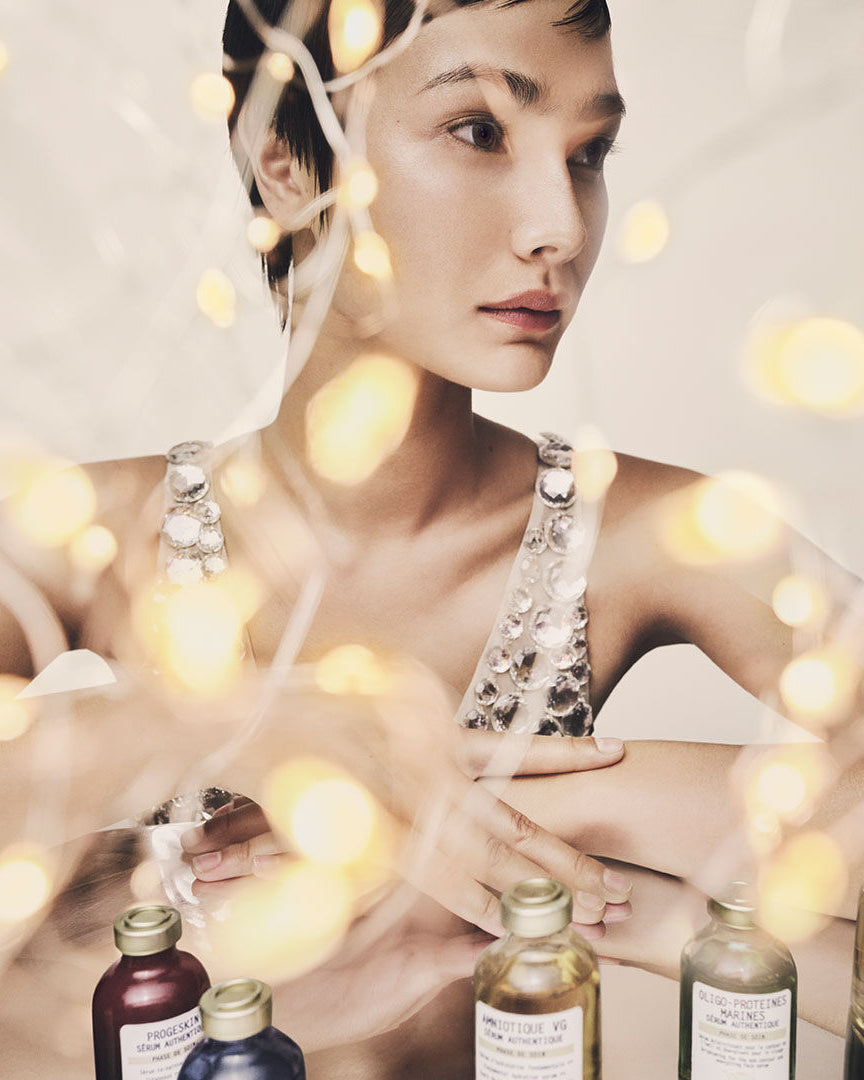 A woman with short dark hair looks off-camera while wearing a silver embellished halter top. In the foreground, several small cosmetic bottles with gold caps are arranged on a surface, partially obscured by warm glowing fairy lights.