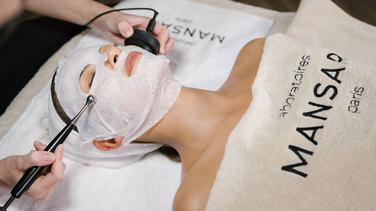 A person lying down with a white facial sheet mask on their face, while another person uses handheld electronic devices on their head and neck. The person is wrapped in a beige towel with the brand name 'MANSA Paris' printed on it.